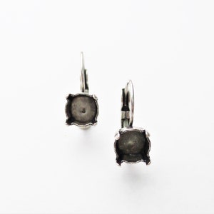 May include: A pair of silver earring settings with a square shape and a small hole in the center. The settings are designed to hold a gemstone or other embellishment.