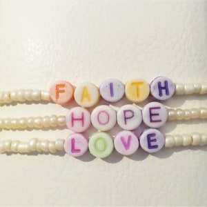 May include: Three beaded bracelets with the words "FAITH", "HOPE", and "LOVE" spelled out in colorful letter beads. The bracelets are made of small, off-white beads and are displayed on a white surface.