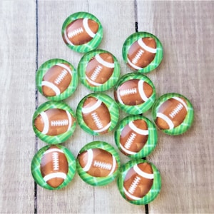 May include: A set of 14 round glass cabochons with a football design on a green grass background. The footballs are brown with white laces.