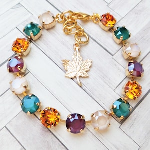 May include: A gold bracelet with a charm of a maple leaf. The bracelet is adorned with colorful crystals in shades of green, purple, orange, and white.