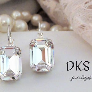 May include: A pair of silver dangle earrings with clear emerald-cut crystal stones. The earrings are on a white surface with the text "DKS jewelrydesigns" in black.