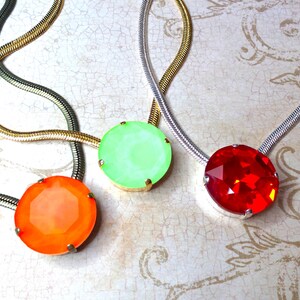 May include: Three necklaces with large, round gemstone pendants in orange, green, and red. The pendants are set in silver-colored metal and hang from snake chains in silver and gold tones. The background has a floral pattern.