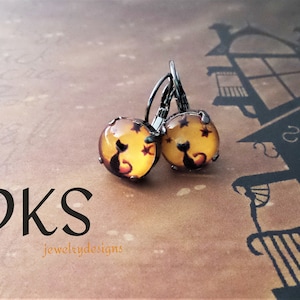 May include: A pair of dangle earrings with a round, orange glass cabochon featuring a black silhouette of a cat with stars. The earrings are silver-toned and have a hook closure. The earrings are on a brown background with the text "DKS jewelrydesigns".