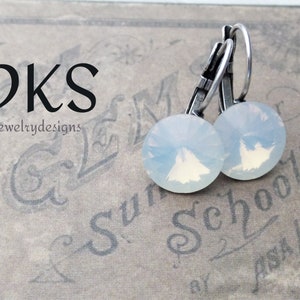 May include: A pair of silver earrings with white opal stones. The earrings have a hook closure. The earrings are on a textured surface with the text "DKS jewelrydesigns" and "Sun Schools".