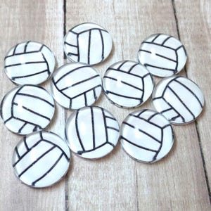May include: A collection of ten round glass cabochons featuring a volleyball design. Each cabochon has a white background with black lines creating the volleyball pattern. The cabochons are arranged on a light brown wooden surface.