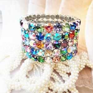 May include: A multi-colored crystal bracelet with a silver metal band. The bracelet is made up of many small, round crystals in various colors, including blue, green, red, orange, and purple. The bracelet is sitting on a white pearl necklace.