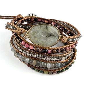 May include: A multi-strand wrap bracelet featuring a large oval-shaped speckled grey stone centrepiece. The bracelet is constructed with brown cord and incorporates various small beads in shades of pink, grey, clear, and dark green throughout.