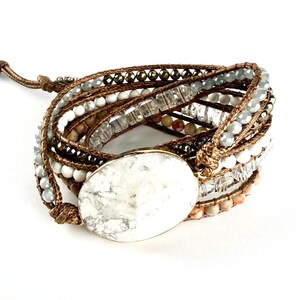 May include: A multi-strand wrap bracelet featuring a large, oval-shaped white stone centrepiece with grey veining. The bracelet is constructed with brown cord and includes various beads, including white, clear, and metallic grey faceted stones.