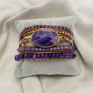 Könnte beinhalten: Wickelarmband mit einem ovalen Amethyststein in einem goldfarbenen Rahmen. Das Armband hat mehrere Stränge aus lila Perlen, goldfarbenen Perlen und braunem Kordel. Das Armband wird auf einem grauen Kissen präsentiert.