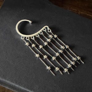 May include: Silver ear cuff featuring a curved top that hooks over the ear. Multiple delicate chains cascade down, each adorned with small star-shaped charms. The jewelry piece is designed to be worn on the ear.