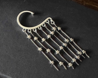 May include: Silver ear cuff featuring a curved top that hooks over the ear. Multiple delicate chains cascade down, each adorned with small star-shaped charms. The jewelry piece is designed to be worn on the ear.