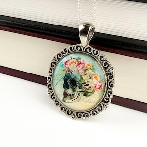 May include: A silver-toned pendant necklace featuring a round, ornate frame. The pendant showcases a skull adorned with flowers and a turquoise eye. The skull is set against a light blue background, with a silver chain.