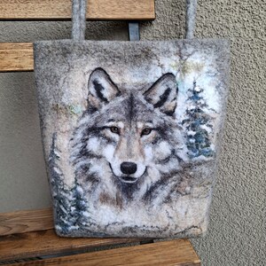 Puede incluir: Bolso tote de fieltro gris con un retrato detallado de un lobo. La cara del lobo est&aacute; centrada, con un fondo de bosque nevado. El bolso tiene dos asas grises y se muestra en un banco de madera.