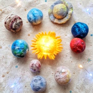 Può includere: Un set di sfere in lana infeltrita che rappresentano un sistema solare, con un sole giallo brillante a punte radianti circondato da otto pianeti multicolori in varie tonalità di blu, rosso, rosa e grigio, disposti su un tessuto a motivi stellati.