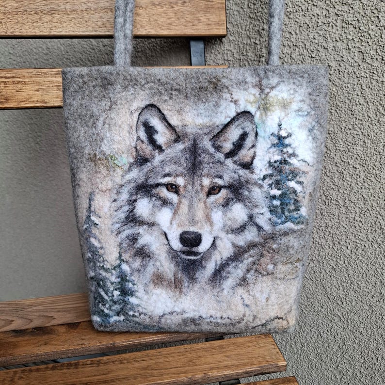 Puede incluir: Bolso tote de fieltro gris con un retrato detallado de un lobo. El lobo tiene pelaje gris y blanco, con una mirada enfocada. El fondo incluye una escena de bosque nevado con &aacute;rboles de hoja perenne. El bolso tiene asas grises.