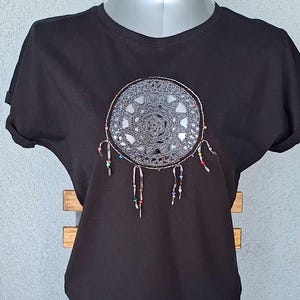 Può includere: Una t-shirt nera a maniche corte con un motivo circolare di acchiappasogni all'uncinetto grigio sul petto. L'acchiappasogni presenta strisce di tessuto pendenti decorate con piccole perline multicolori che scendono dal bordo inferiore.