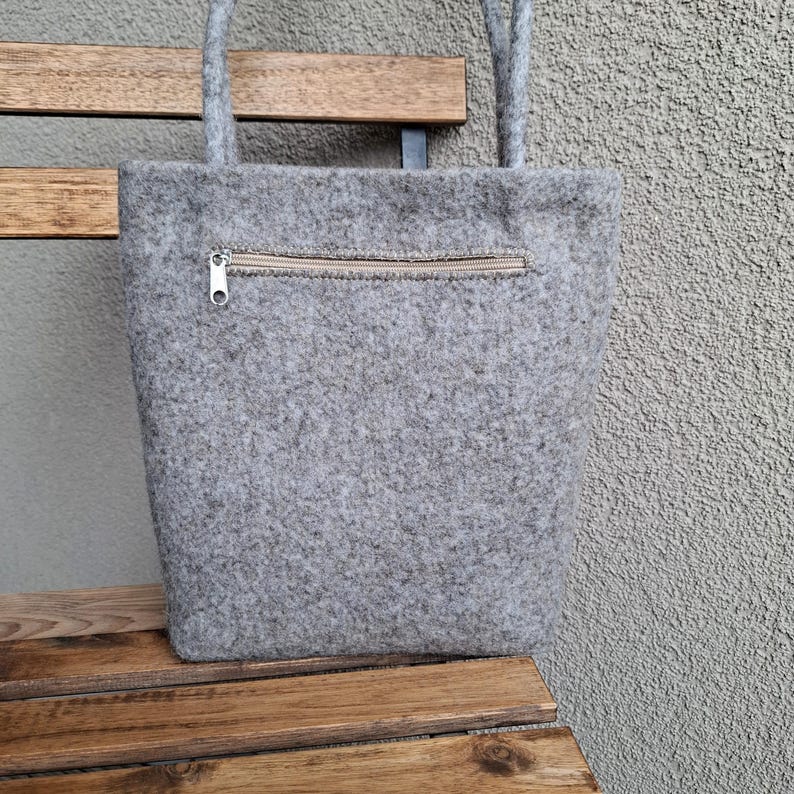 Puede incluir: Un bolso tote de fieltro gris con un bolsillo con cremallera en la parte delantera. El bolso tiene dos asas y est&aacute; sobre un banco de madera. La cremallera es de color beige claro.