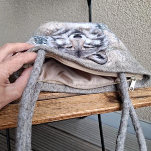 Puede incluir: Un bolso de fieltro gris con un dise&ntilde;o de cara de lobo. El bolso tiene una cremallera en la parte superior y asas de cuerda gris. El interior es de color beige claro. La cara del lobo est&aacute; detallada con tonos de gris y blanco.