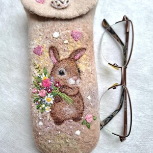 Könnte beinhalten: Ein beiges Brillenetui aus gefilzter Wolle mit einem gestickten braunen Hasen, der einen bunten Blumenstrauß hält. Das Etui hat einen Klappverschluss mit einem dekorativen Perlenknopf und ist mit kleinen rosa Stickherzen und Perlen verziert.