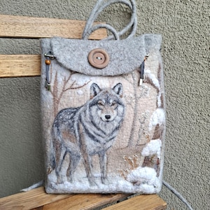 May include: A light gray felted backpack featuring a needle-felted wolf standing in a snowy forest scene. The bag has a flap closure secured by a large wooden button, two top handles, and gray cord drawstrings with metallic silver end caps.