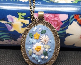 May include: A blue fabric pendant necklace with a bronze oval frame. The pendant features embroidered white, pink, and yellow flowers with green leaves.