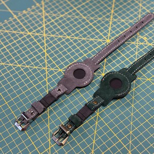 Può includere: Due cinturini per orologio in pelle con supporti circolari per un dispositivo. Un cinturino è grigio con cuciture bianche e fibbia argentata, l'altro è verde scuro con cuciture bianche e fibbia color bronzo. Entrambi hanno un foro circolare.