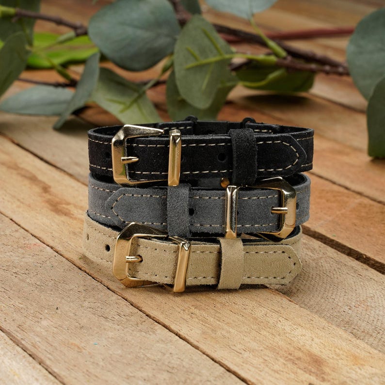 May include: A stack of three suede bracelets in black, gray, and beige. Each bracelet features a gold-toned metal buckle, a matching metal keeper, and visible stitching along the edges of the strap, creating a layered and textured appearance.
