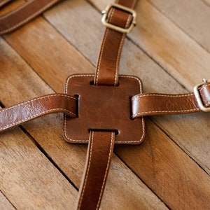 Può includere: Un cinturino in pelle marrone con una toppa quadrata in pelle e fibbie metalliche. Il cinturino è cucito con filo bianco ed è progettato per essere utilizzato come cinturino per fotocamera.