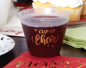 May include: A clear plastic cup filled with a dark red drink. The cup has gold lettering that says "* cup of cheer *" on the side. The cup is sitting on a red napkin with gold lettering that says "merry and bright".