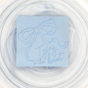 May include: A light blue napkin with a white embossed bow and the text "Tying the knot" on a white marble plate.