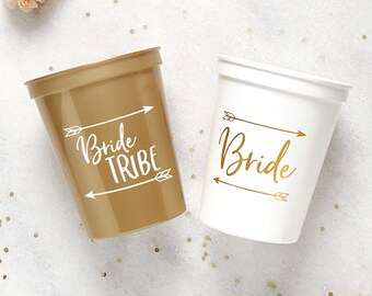 May include: Two plastic cups with gold glitter accents. One cup is gold and says "Bride Tribe" in white lettering. The other cup is white and says "Bride" in gold lettering.