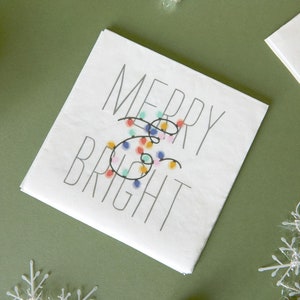 Op de afbeelding: Witte kaart met de tekst "Merry & Bright" in zwarte letters. Het ampersand is versierd met een reeks gekleurde lampjes.