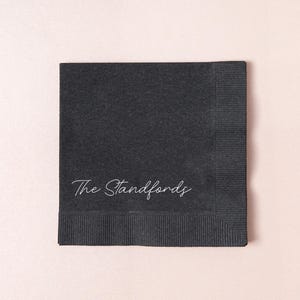 May include: A square, charcoal-colored paper napkin with the text "The Standfords" in a delicate, white script. The napkin has a textured surface and a ribbed border along one edge. Ideal for events.