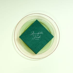 May include: A top-down view of a layered place setting. A dark green square napkin is centered on a clear green plate, which rests on a larger, clear plate with a gold rim. The napkin has the text "Annabelle and Joseph 5.15.2035" in white script.