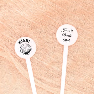 May include: Two white plastic drink stirrers with round tops. One stirrer has a black and white seashell design with the word "MIAMI" printed above it. The other stirrer has the text "Jenn's Bach Club" printed in black.