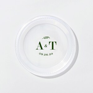 May include: A clear, round plastic plate featuring a dark green printed design in the center. The design includes a small leafy sprig above the letters A & T, with the date 09.26.24 printed in a curved dark green font below the letters.