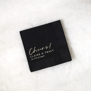 May include: A black square napkin with gold text that reads "Cheers!" followed by "CLAIRE & TRACY" and "the 27th of august". The napkin is on a white marble surface. The napkin is likely for a wedding or special event.