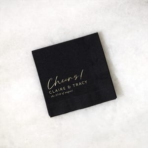 May include: Black square cocktail napkin with gold script text that reads "Cheers!" followed by "CLAIRE & TRACY" and "the 27th of august". The napkin is on a white marble surface.