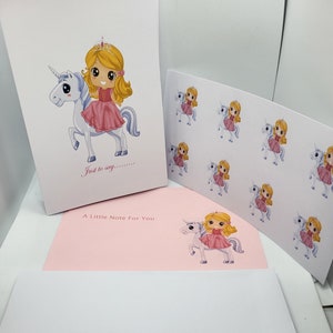 May include: A stationery set featuring a white greeting card with a cartoon girl in a pink dress riding a white unicorn and the text Just to say. Includes a sheet of matching stickers, a light pink note card, and a white envelope with the same graphic.