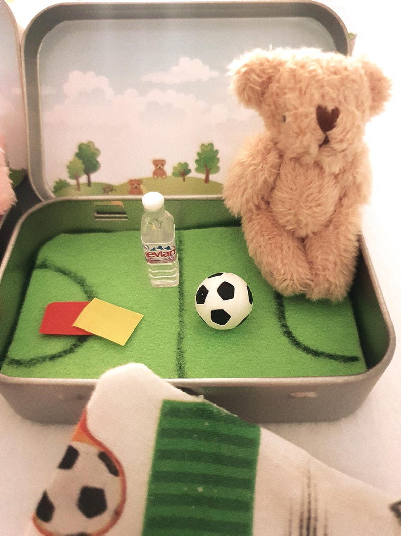 Tiny 8cm teddy in pocket tin laying on green felt football pitch with football 100 percent cotton blanket teddy has a football yellow and red warning cards and a football. Personalisation is optional