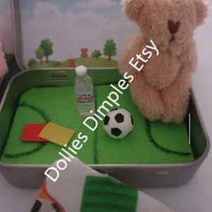 Tiny 8cm teddy in pocket tin laying on green felt football pitch with football 100 percent cotton blanket teddy has a football yellow and red warning cards and a football. Personalisation is optional