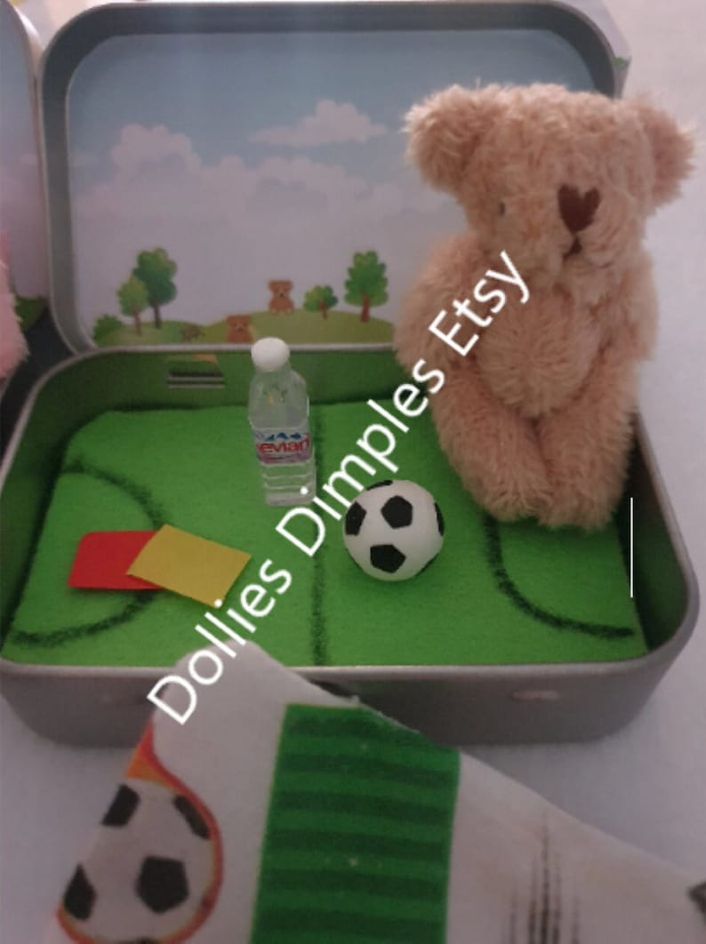 Tiny 8cm teddy in pocket tin laying on green felt football pitch with football 100 percent cotton blanket teddy has a football yellow and red warning cards and a football. Personalisation is optional