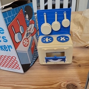Puede incluir: Un juego de cocina de juguete vintage en azul y crema. El juego incluye una estufa con dos quemadores, un protector contra salpicaduras azul con tres sartenes colgantes y una caja con una ilustración retro de una mujer cocinando.