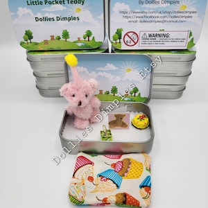 Puede incluir: Un oso de peluche rosa con un sombrero amarillo se sienta en una lata de plata con una almohada de cupcake y otros pequeños accesorios. La lata tiene una etiqueta que dice "Little Pocket Teddy Dollies Dimples".