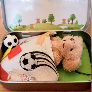 Tiny 8cm teddy in pocket tin laying on green felt football pitch with football 100 percent cotton blanket teddy has a football yellow and red warning cards and a football. Personalisation is optional