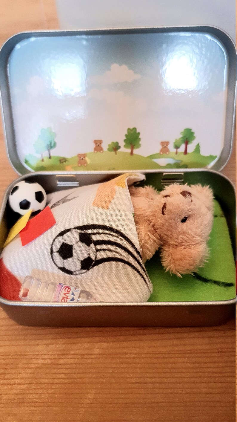 Tiny 8cm teddy in pocket tin laying on green felt football pitch with football 100 percent cotton blanket teddy has a football yellow and red warning cards and a football. Personalisation is optional