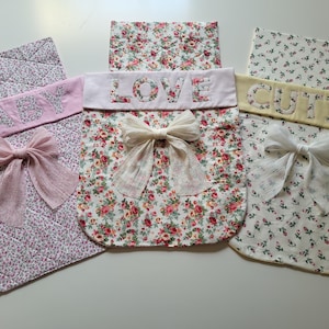 Puede incluir: Tres bolsas rectangulares de tela acolchada con estampados florales en rosa, rojo y verde. Cada bolsa tiene una banda de color sólido con el texto bordado BABY, LOVE o CUTIE, y está decorada con un lazo de tela transparente en el centro.