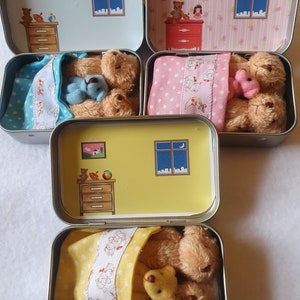 May include: Several small, open metal tins with interior illustrations of bedrooms. Each tin contains a teddy bear and a smaller bear, nestled under a patterned blanket. The tin interiors feature a night sky scene and furniture.