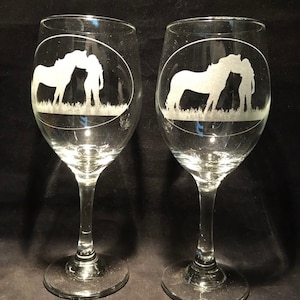 Op de afbeelding: Twee heldere glazen wijnglazen met gegraveerde ontwerpen van een vrouw en een paard. Het ontwerp is ovaal en het paard kust de vrouw op de wang.