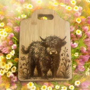 May include: A wooden cutting board with a handle, featuring a detailed etched image of a Highland cow. The cow is depicted in a field of grass and flowers. The board is surrounded by a colorful floral background.